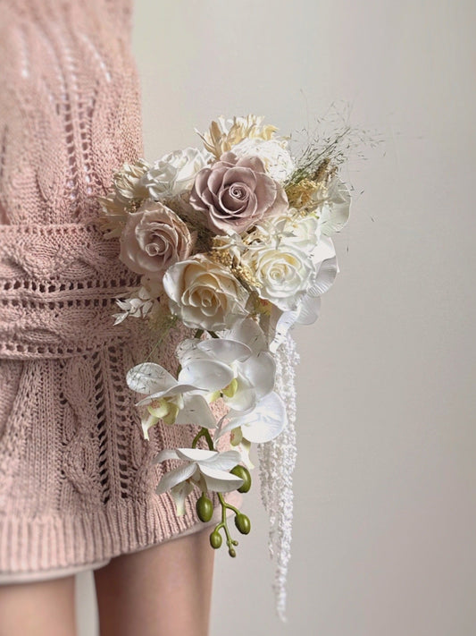 Soft pastel hues preserved roses bridal bouquet in teardrop style, completed with a faux phalaenopsis orchid. Perfect for elegant brides seeking a refined and timeless floral arrangement.
专为优雅新娘打造的经典永恒花艺设计。