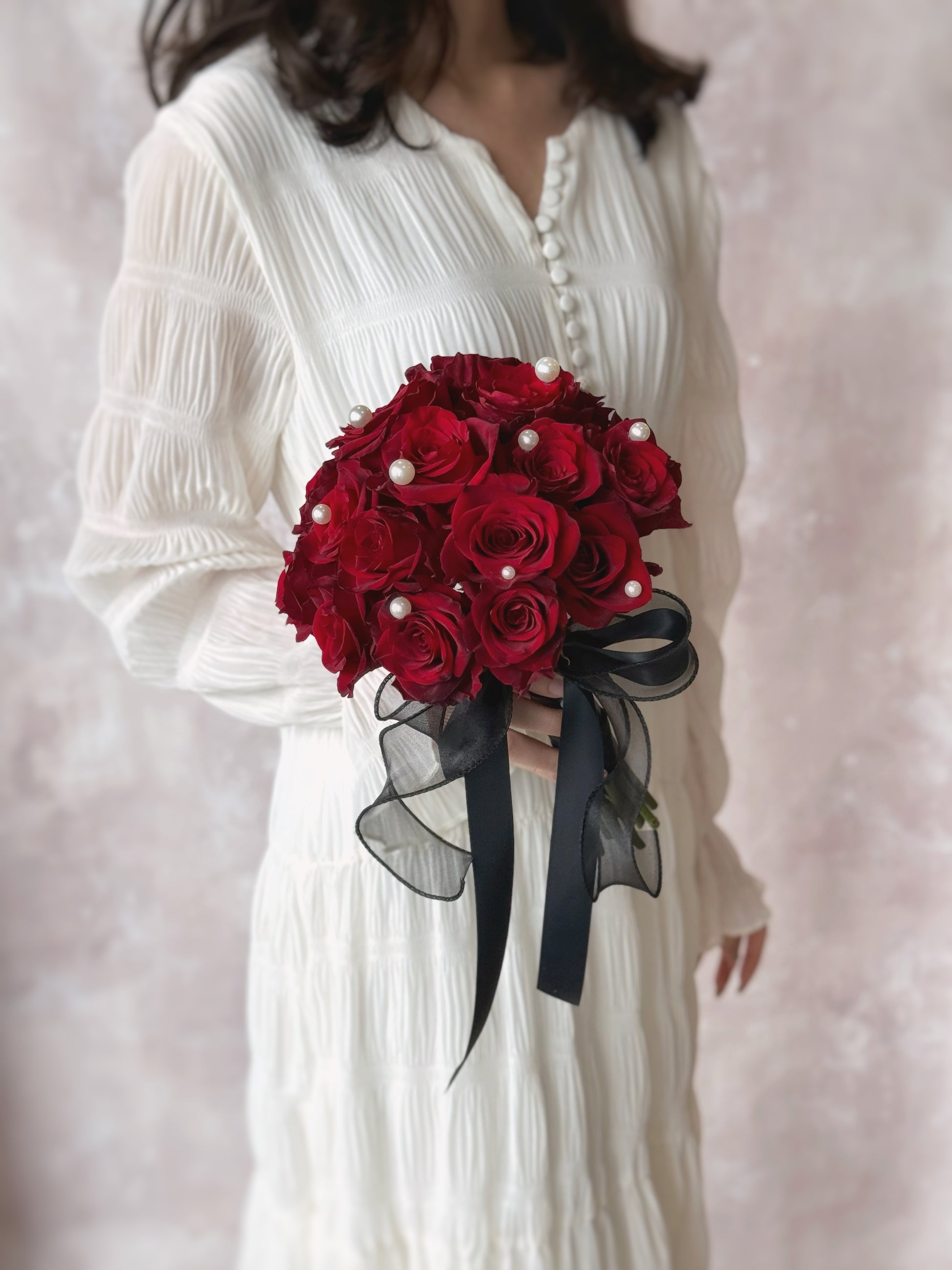 Zoomed out view of a model in white dress holding a bold maroon rose bouquet, adorned with pearls and a black ribbon for a striking and elegant look.
传统与现代结合的新娘捧花,东方风格装饰尽显高雅