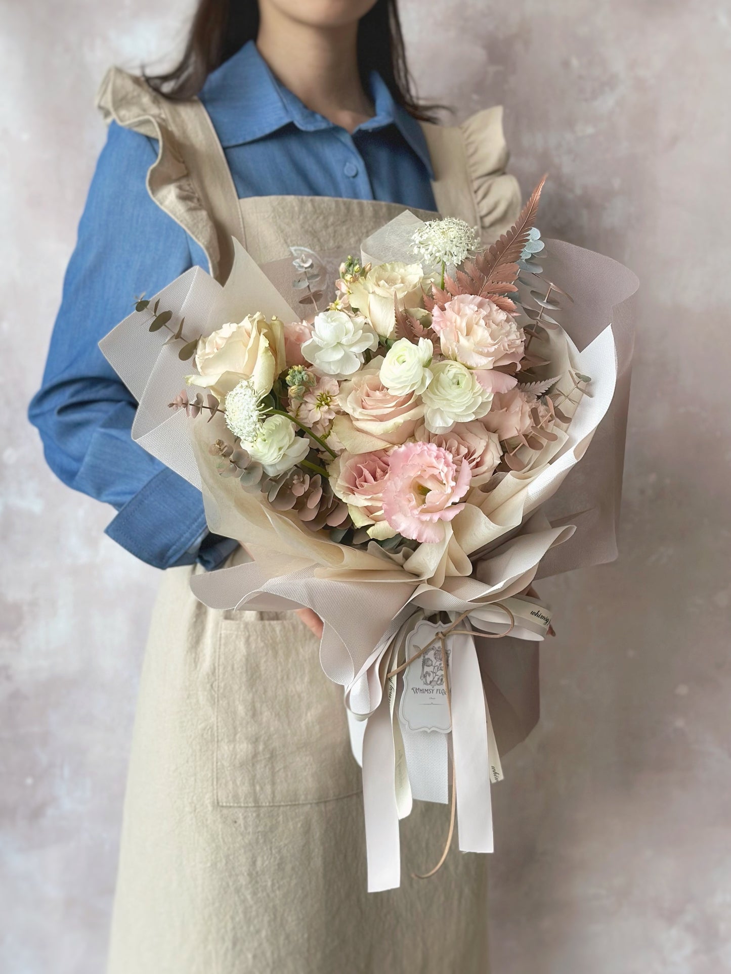 A model holding a grand size pastel brown-pink bouquet with quicksand roses and painted foliage, styled for a dreamy neutral-toned look.
模特手捧一束棕粉色系花束,搭配柔和的玫瑰与喷染叶材,整体风格梦幻而自然。