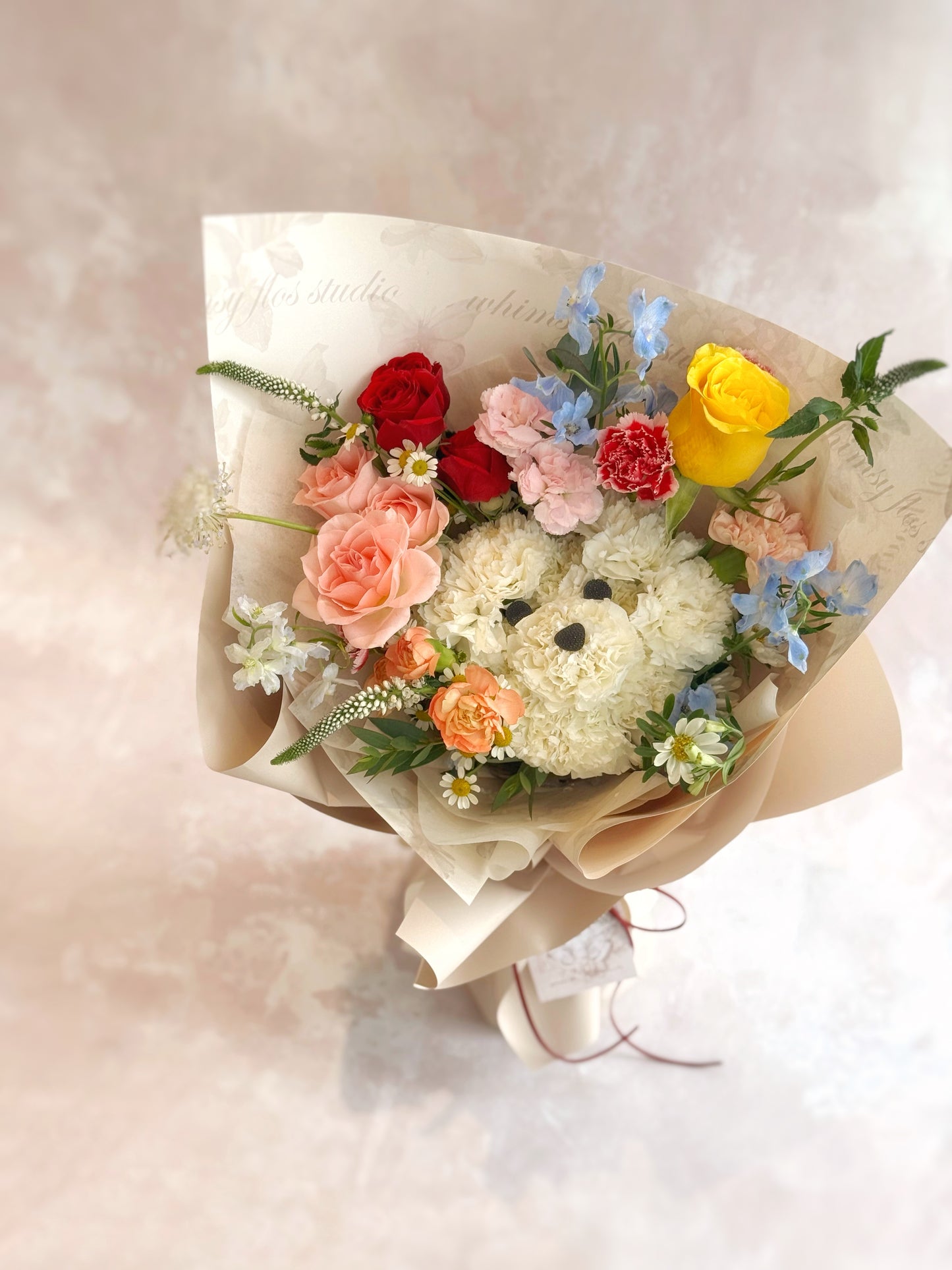 Bouquet of flowers with a teddy bear-shaped center on a light background. Top-down view of bouquet showing the white puppy face at center, surrounded by vibrant blooms in a round, cheerful arrangement.
俯视图呈现花束中心的白色小狗造型,外围环绕多彩花材,整体造型圆润且欢乐。