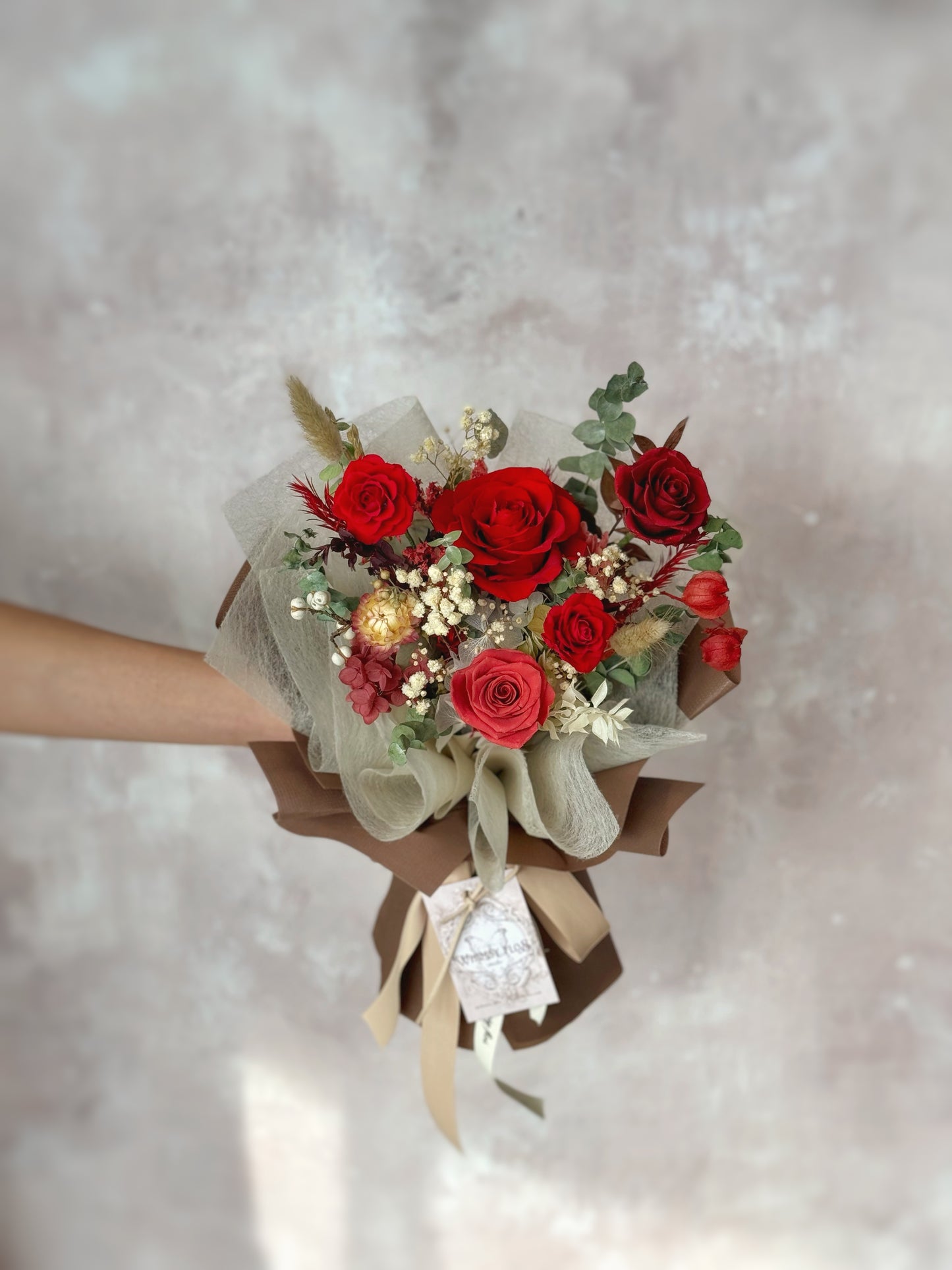 A close-up view of the Crimson Dusk bouquet, highlighting the preserved roses and delicate florals wrapped in earthy brown for a refined, rustic touch.
“Crimson Dusk”花束的特写,突出了永生玫瑰和精致的花卉,包裹在大地棕色中,展现了精致而质朴的风格。