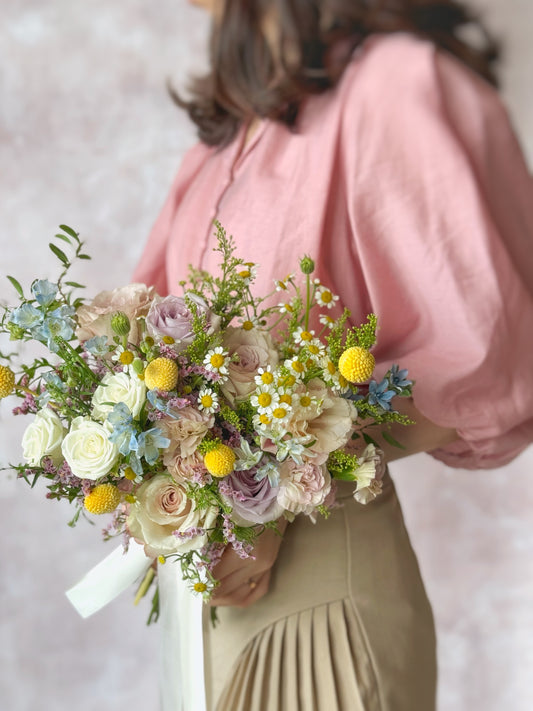 Bride holding Dorothea fresh flowers bridal bouquet, a whimsical pastel roses bouquet in shades of lilac, green, yellow, blue, and pink for a fairytale-inspired look.