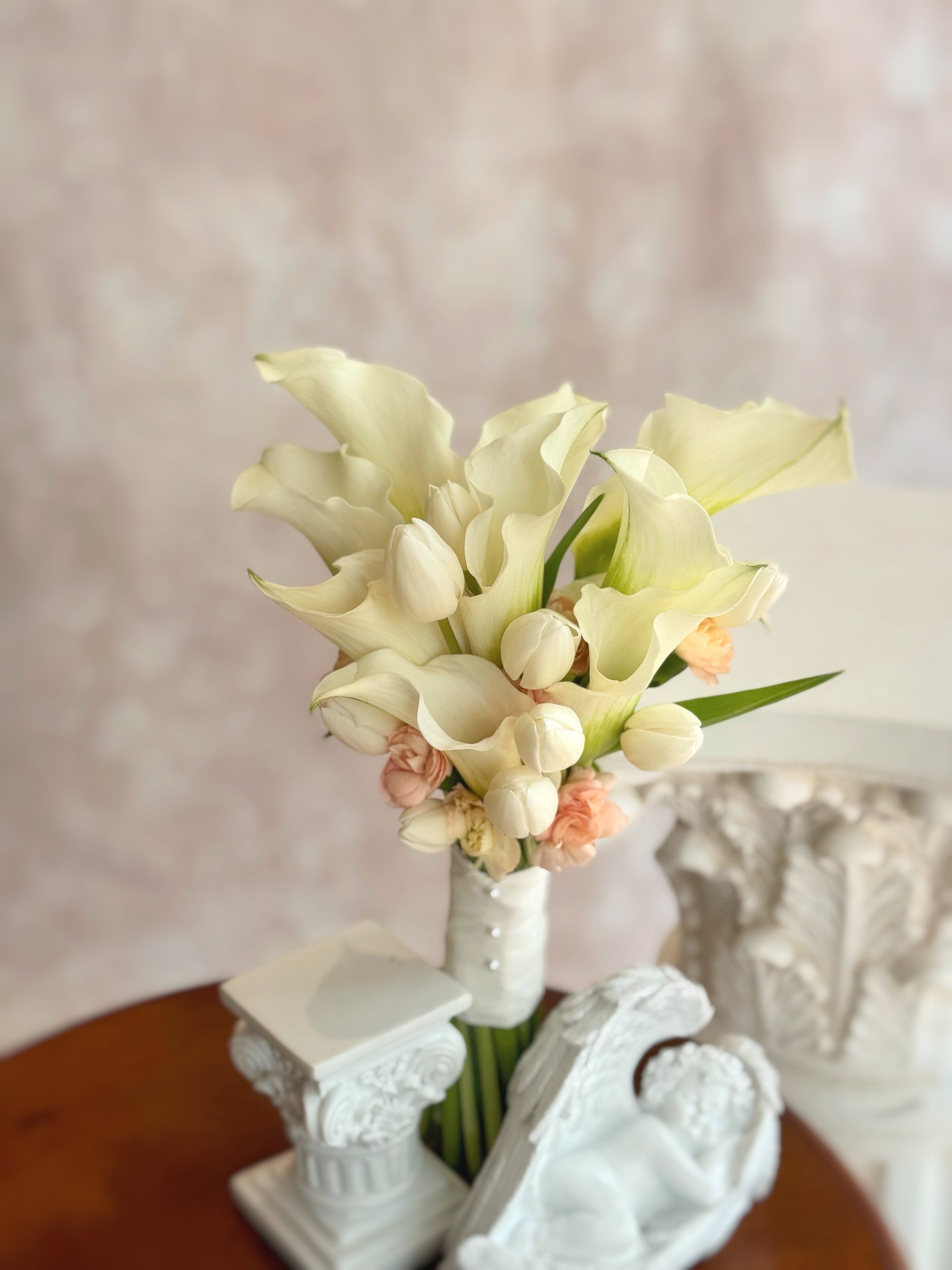 Front view of a minimalist bridal bouquet featuring sleek white calla lilies, soft tulips, and warm nude peach spray carnations.
正面展示简约新娘花束,结合优雅白色马蹄莲、柔和郁金香及暖色调裸粉色小康乃馨。