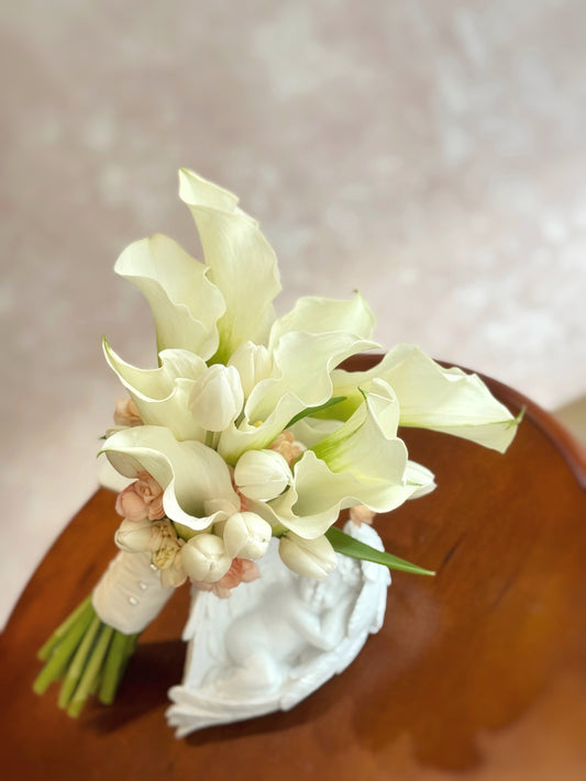Minimalist calla lilies and tulips bridal bouquet arranged on a table, radiating fresh elegance and understated beauty perfect for a wedding setting.
简约白色马蹄莲郁金香新娘捧花摆放于桌面,散发清新优雅与低调美感,适合婚礼氛围。