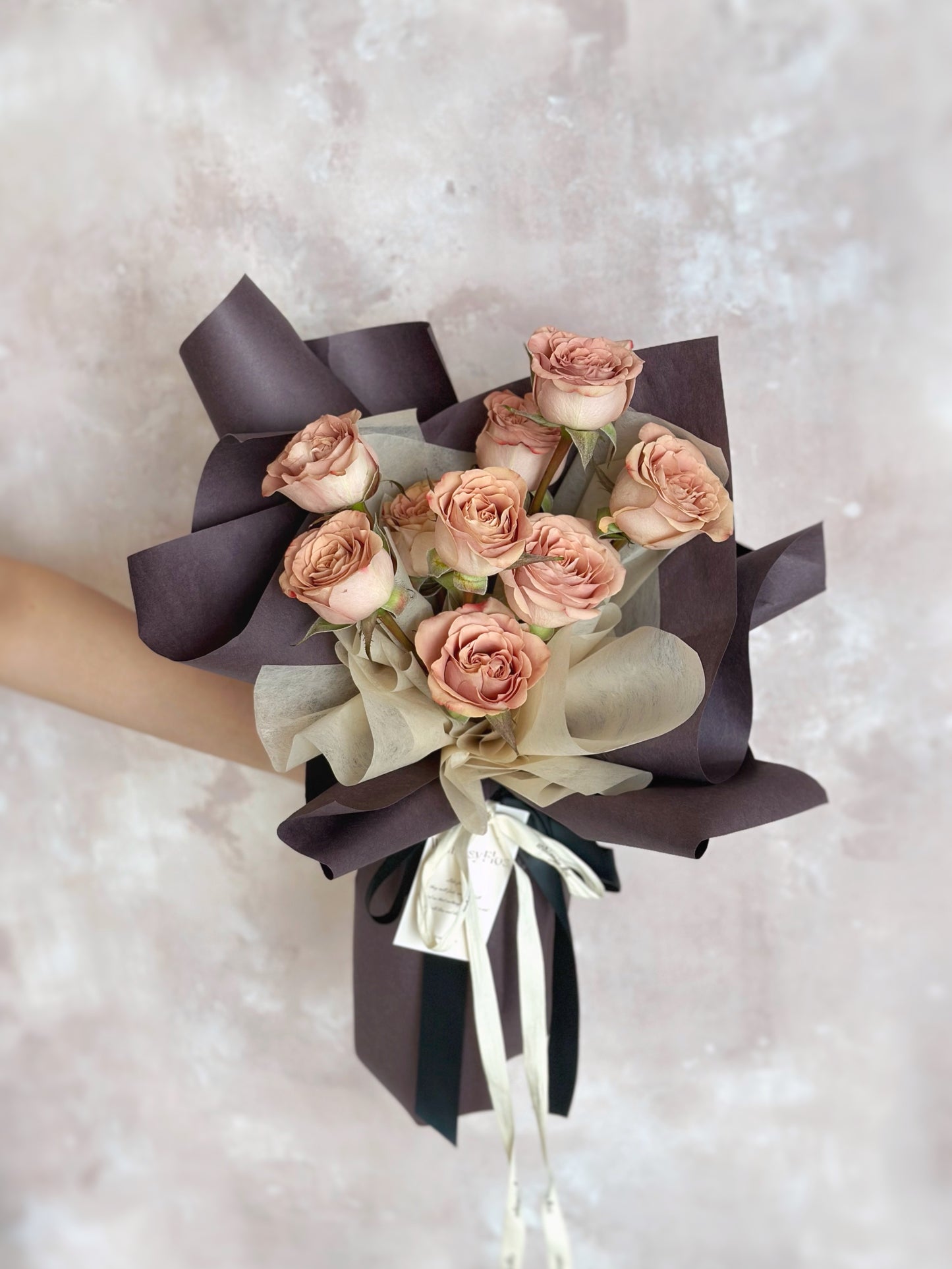 Bouquet held at arm’s length with only hands visible, focusing on the cappuccino roses and sculptural deep mauve wrap.
手臂前伸持花,镜头聚焦卡布奇诺玫瑰与深紫包裹,突显花束质感与格调。