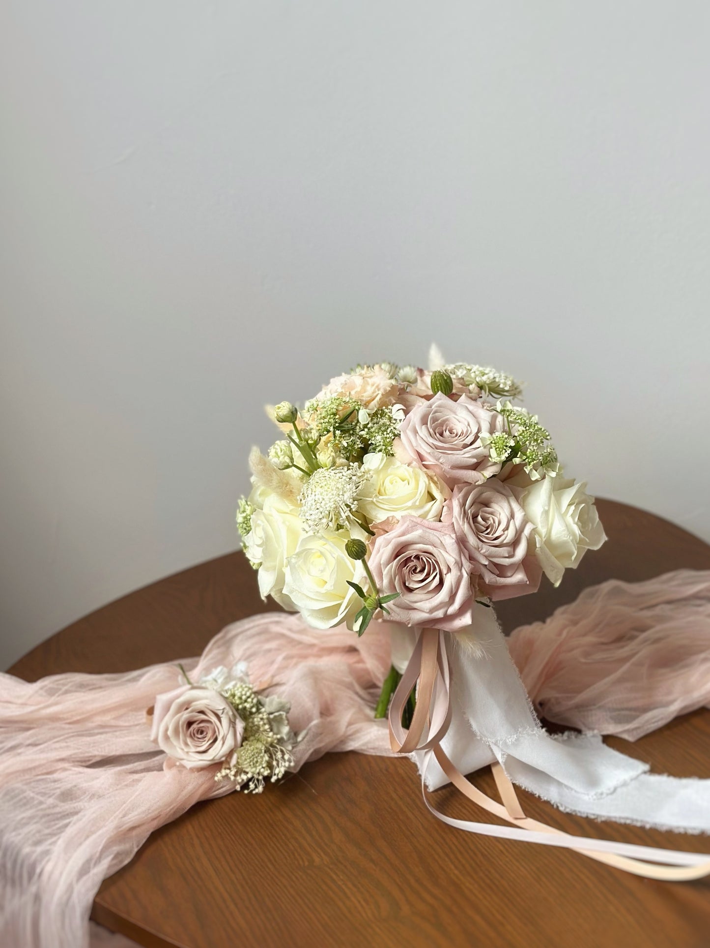 A round bridal bouquet of nude blush Menta, Quicksand roses and cream roses arranged with varying heights for depth and elegance, displayed on a wooden table.
裸粉色的白玫瑰,曼塔玫瑰和流沙玫瑰组成的圆形新娘捧花,以不同高度排列,增添层次感与优雅气息,摆放在木桌上。