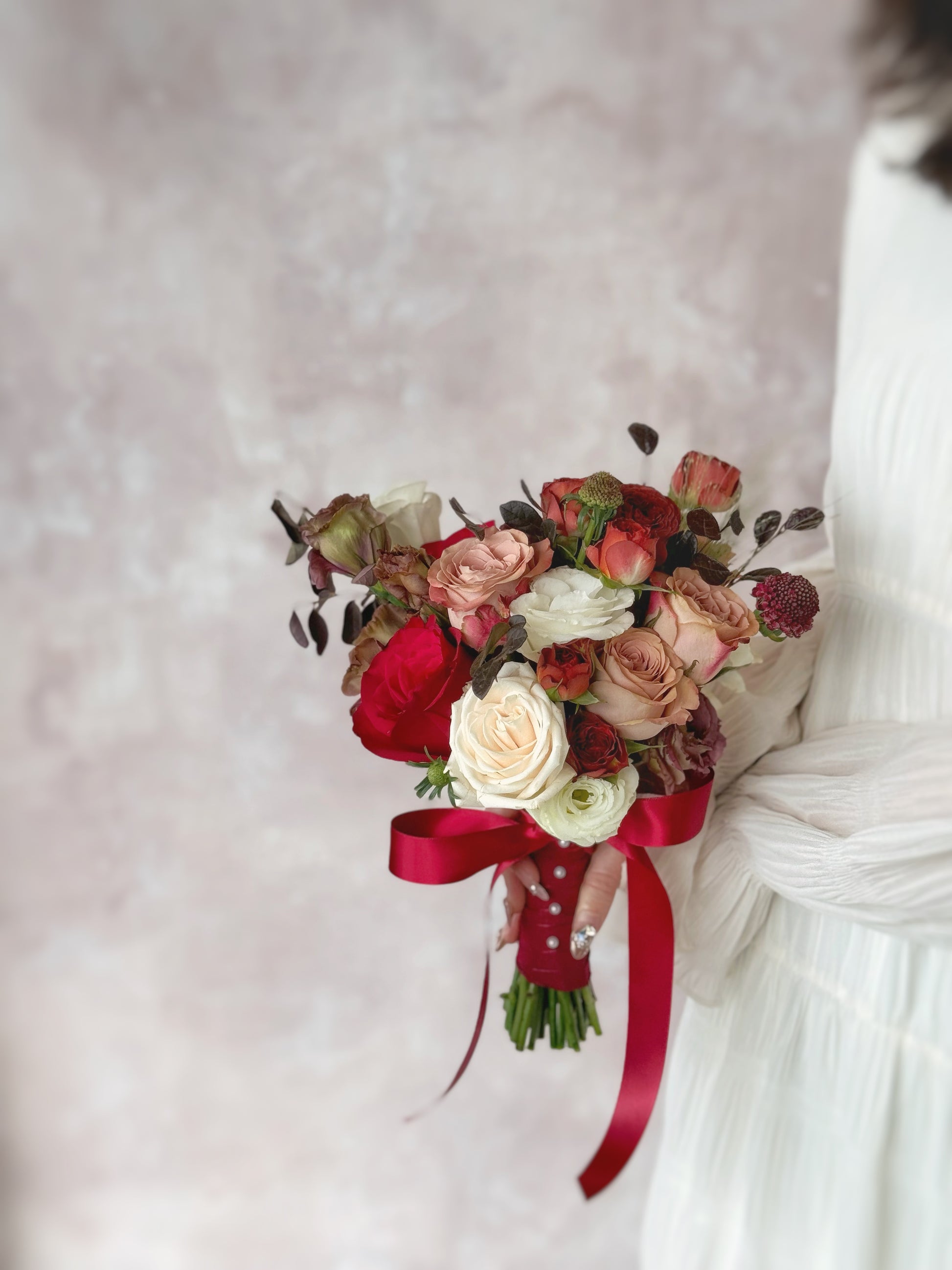 A sophisticated and unique bridal bouquet, featuring rich mauve and maroon tones with delicate blush and white flowers, held by a model at the side
这款捧花的浓郁深色调,与新娘的白色婚纱形成鲜明对比,展现大胆个性