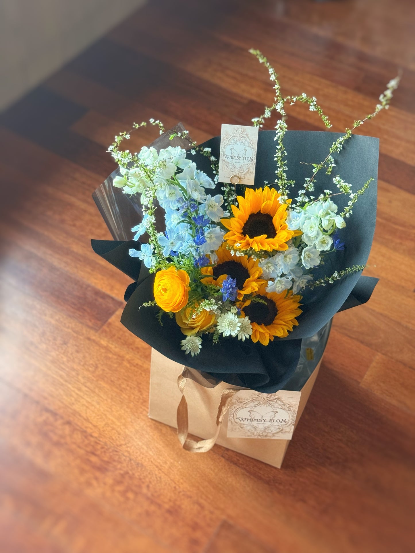 Top-down view showing bold color blocking between the golden sunflowers and the surrounding blue blooms, framed by matte black wrapping.
俯视图呈现黄蓝花材色块拼接效果,整体包裹以哑黑色纸张收边,简约中见张力。