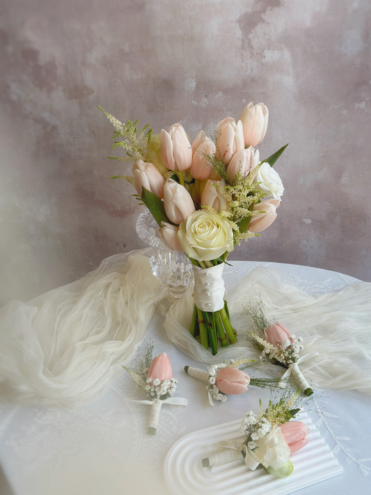 Photo of Korean style peachy pink tulip bridal bouquet on a table, together with  delicate corsages featuring a single peachy tulip and white floral accents, tied with an elegant silk ribbon.
精致韩式蜜桃粉郁金香新娘捧花和与胸花,蜜桃色郁金香搭配白色花朵点缀,丝带装饰更显高贵雅致。