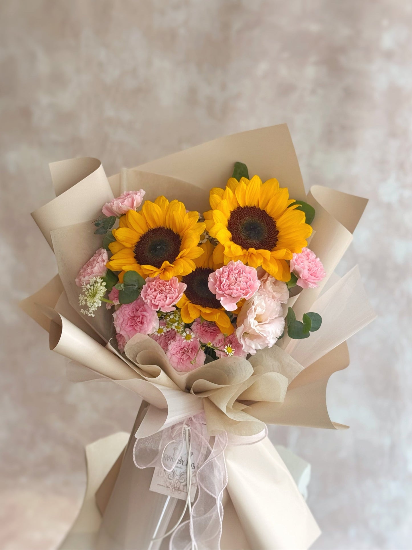 Front view of a hand-tied graduation bouquet with bright sunflowers and soft pink carnations, accented by fillers and greenery.
正面展示手绑毕业季花束,亮黄色向日葵与柔粉康乃馨交织,搭配小配花与绿叶