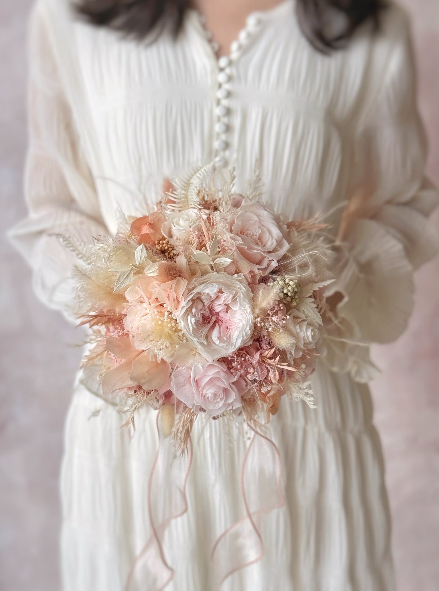Tea rose preserved flowers round shaped bridal bouquet holding in the middle view. A bride holding the preserved flower bouquet, its round shape making it a timeless and classic bridal choice.
新娘手持蜜桃粉白色系温柔永生花捧花,圆形设计让其成为永不过时的经典之选。