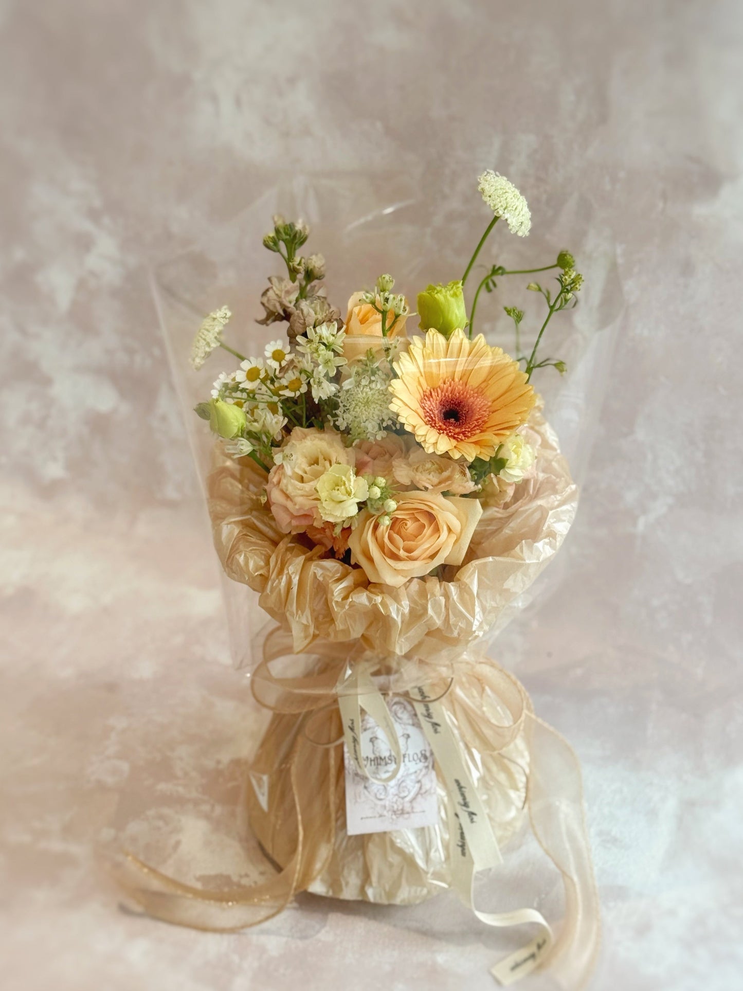 Front view of a hand-tied bouquet featuring fresh florals in peach, cream, khaki, and light brown tones, wrapped in layered textured paper and a sheer wrap.
正面展示手绑花束,由蜜桃、奶油、卡其与浅棕色调花材组成,搭配多层纹理纸与透明轻盈外层包裹。