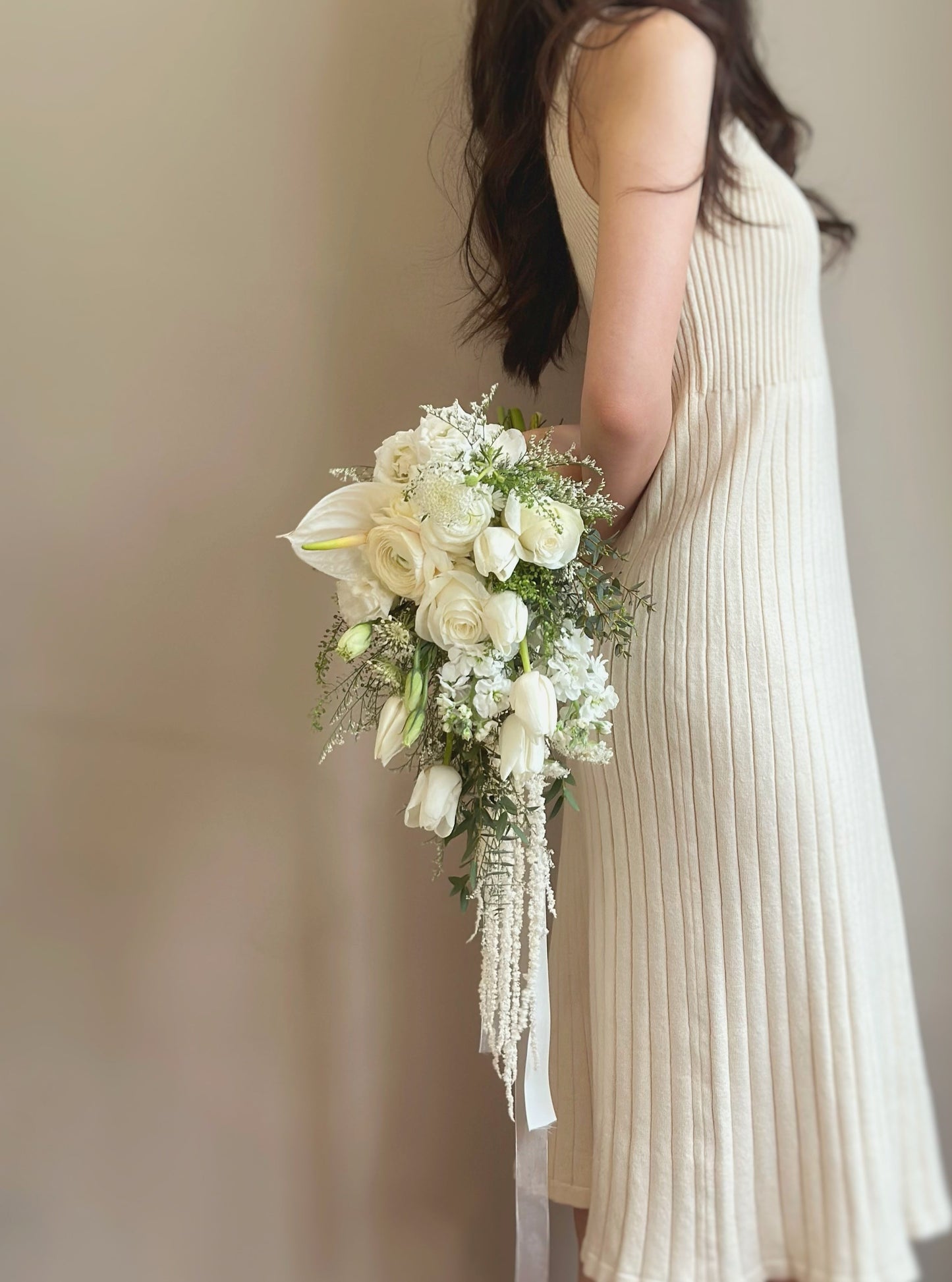 A bride elegantly holding a cascading teardrop bouquet, where white tulips and soft greenery flow gracefully to create a natural and sophisticated look. The bouquet’s elongated silhouette adds an element of movement and charm, making it a captivating choice for brides who love a fairytale aesthetic.
新娘优雅地手持一款垂坠式水滴形捧花,白色郁金香与柔和绿植轻盈流动,营造出自然且高贵的氛围。修长的花束轮廓增添了动感和迷人魅力,非常适合喜爱仙境风格的新娘。