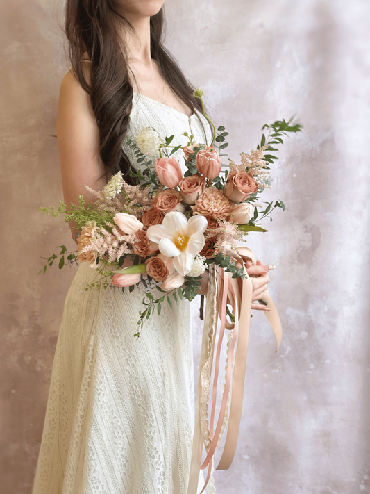 A bride holding a whimsical free-form bouquet with airy greenery and soft blush tulips, cappuccino roses, astilbe, carnation with assorted flowers and foliage, exuding a natural elegance.
新娘手持随性自由形态捧花,轻盈绿叶与浅粉花朵相映,散发自然优雅气息。