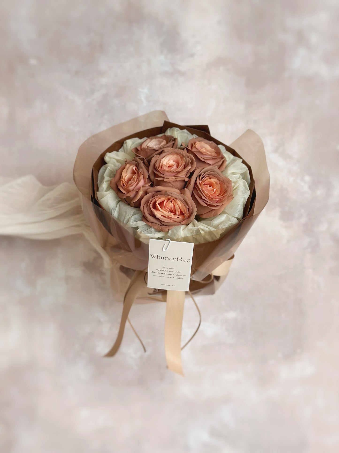 Bouquet held at arm’s length with only hands visible, focusing on the softly tinted roses and layered vintage wrapping.
手臂前伸持花,仅露出手部与花面,玫瑰柔和色泽与复古包裹成为画面焦点