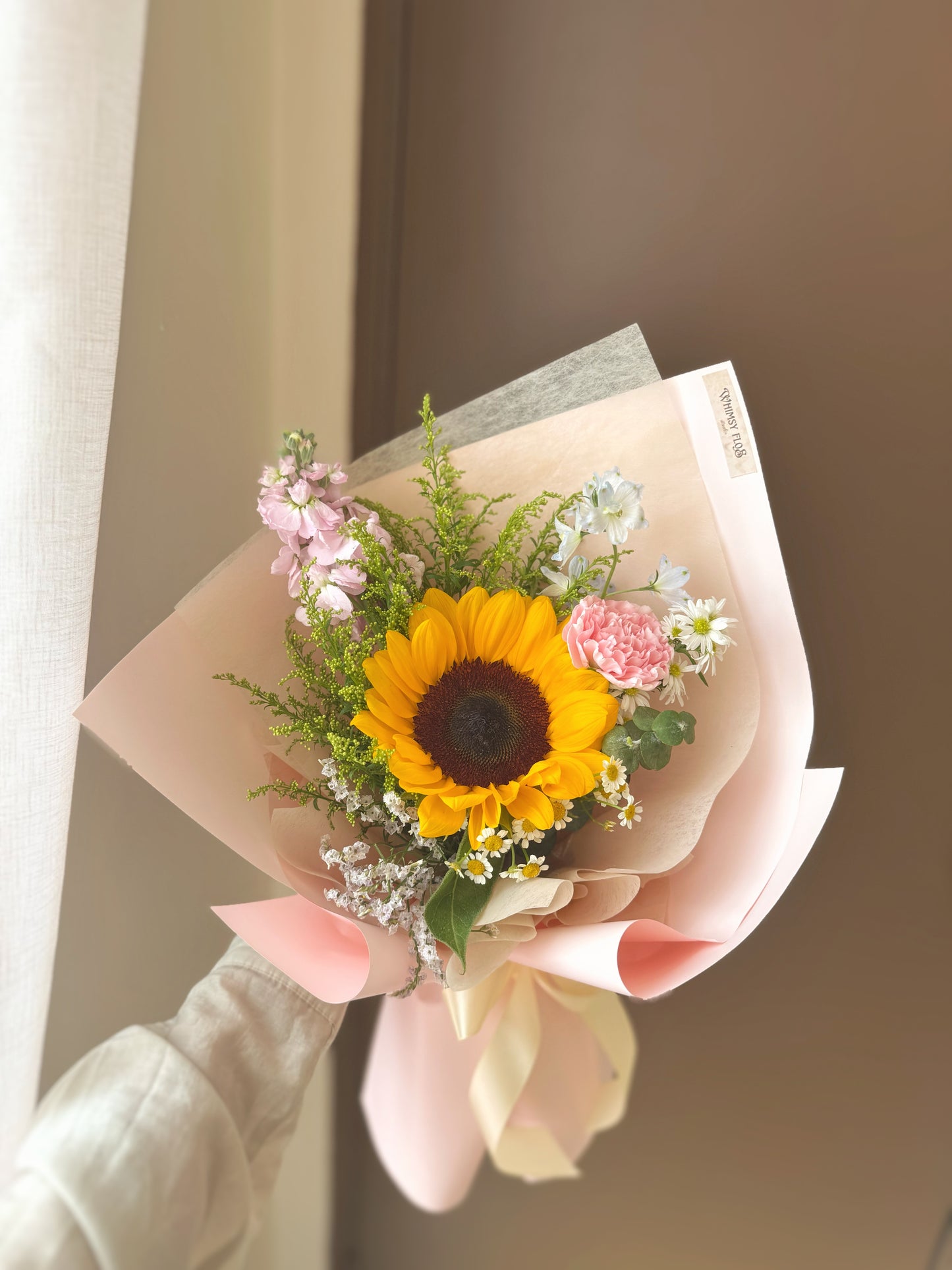 Bouquet held outward at arm’s length, with just the single sunflower and pastel mix in focus — a bright and tender visual moment.
花束由手臂前伸持握,仅呈现花面与手部,向日葵与柔色小花交相辉映,充满温柔明亮的画面感。