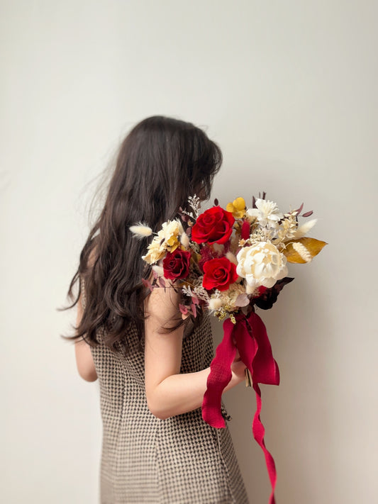 A bride holding the Amber preserved flowers bridal bouquet, its warm autumnal red and mustard yellow palette complementing the season’s romantic charm.
新娘手持Amber捧花，温暖秋色与浪漫气息完美相融。
