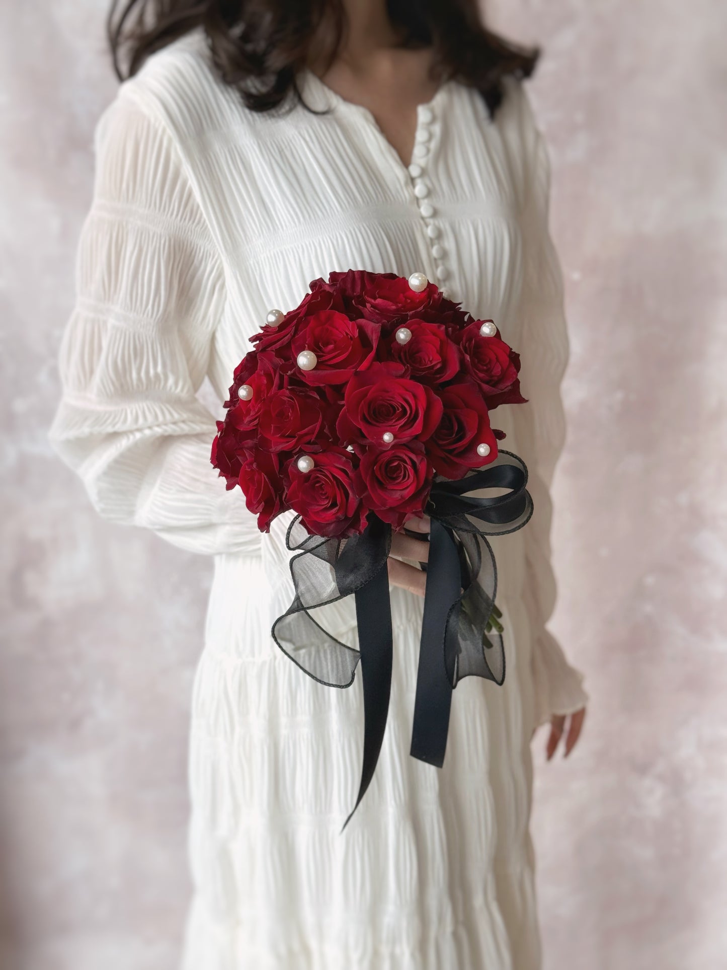 Zoomed out view of a model in white dress holding a bold maroon rose bouquet, adorned with pearls and a black ribbon for a striking and elegant look.
传统与现代结合的新娘捧花，东方风格装饰尽显高雅
