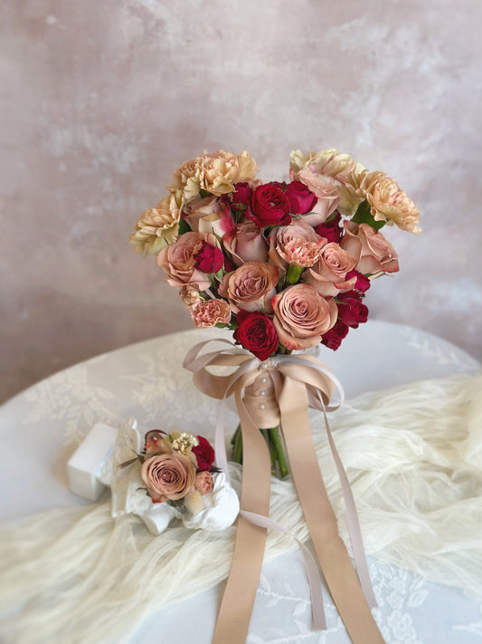 A set of lovely vintage themed fresh flowers bridal bouquet and groom’s boutonnière, featuring cappuccino roses, carnations, spray carnations, ranunculus and etc. A heart-shaped bridal bouquet featuring cappuccino pink and maroon red blooms, carefully arranged to symbolize love and devotion, displayed elegantly on a table.
心形新娘捧花，以卡布奇诺粉色和深酒红色花朵精心排列，象征爱与承诺，优雅地摆放在木桌上。
