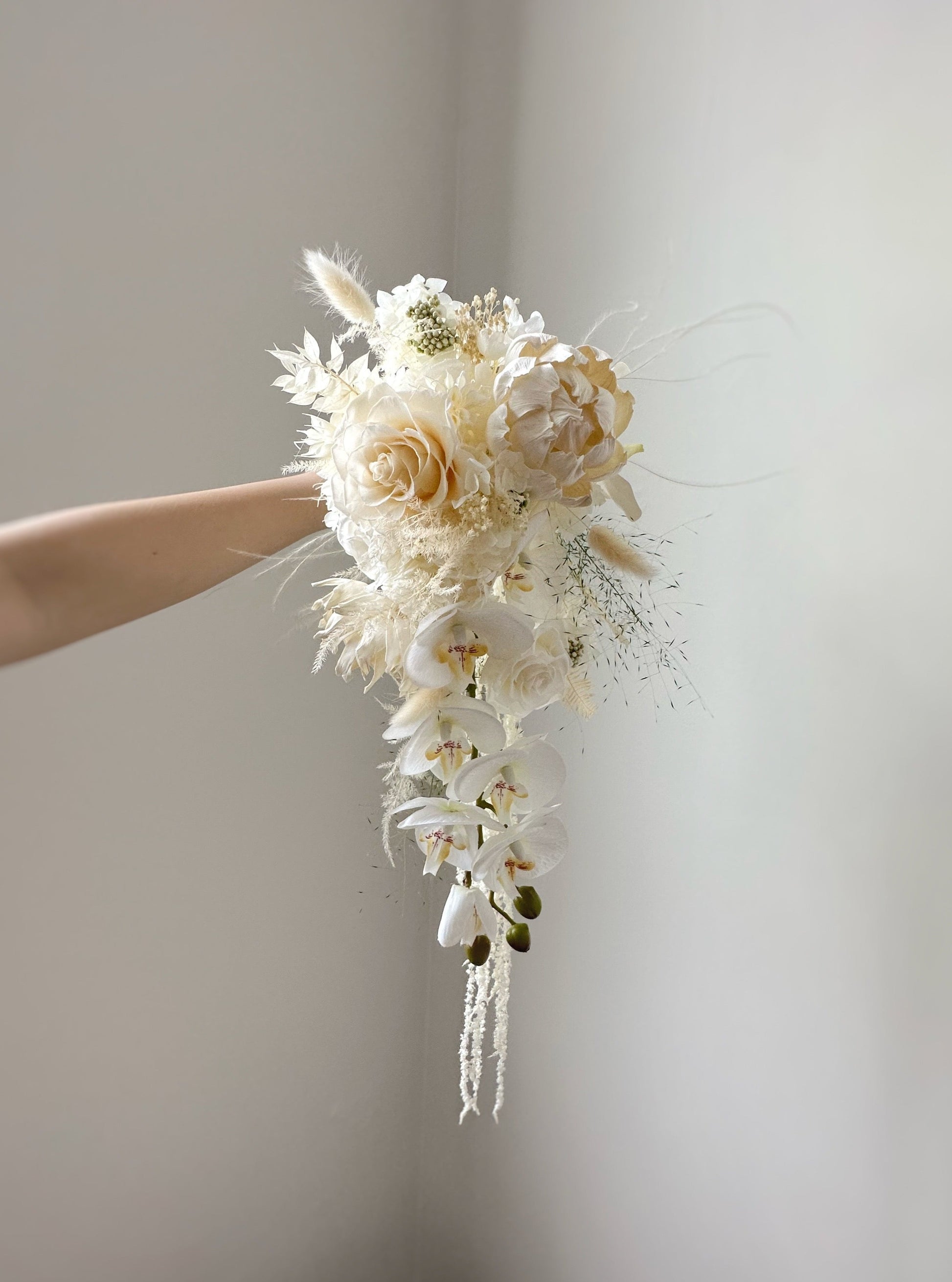Cascading/ teardrop shaped preserved flowers bridal bouquet, creamy white in color, featuring preserved roses, sola flowers and other fillers. A delicate blend of ivory and pastel florals, highlighting intricate textures and soft hues.
象牙白与粉彩花材的细腻搭配，展现精致的花卉质感与柔和色调。