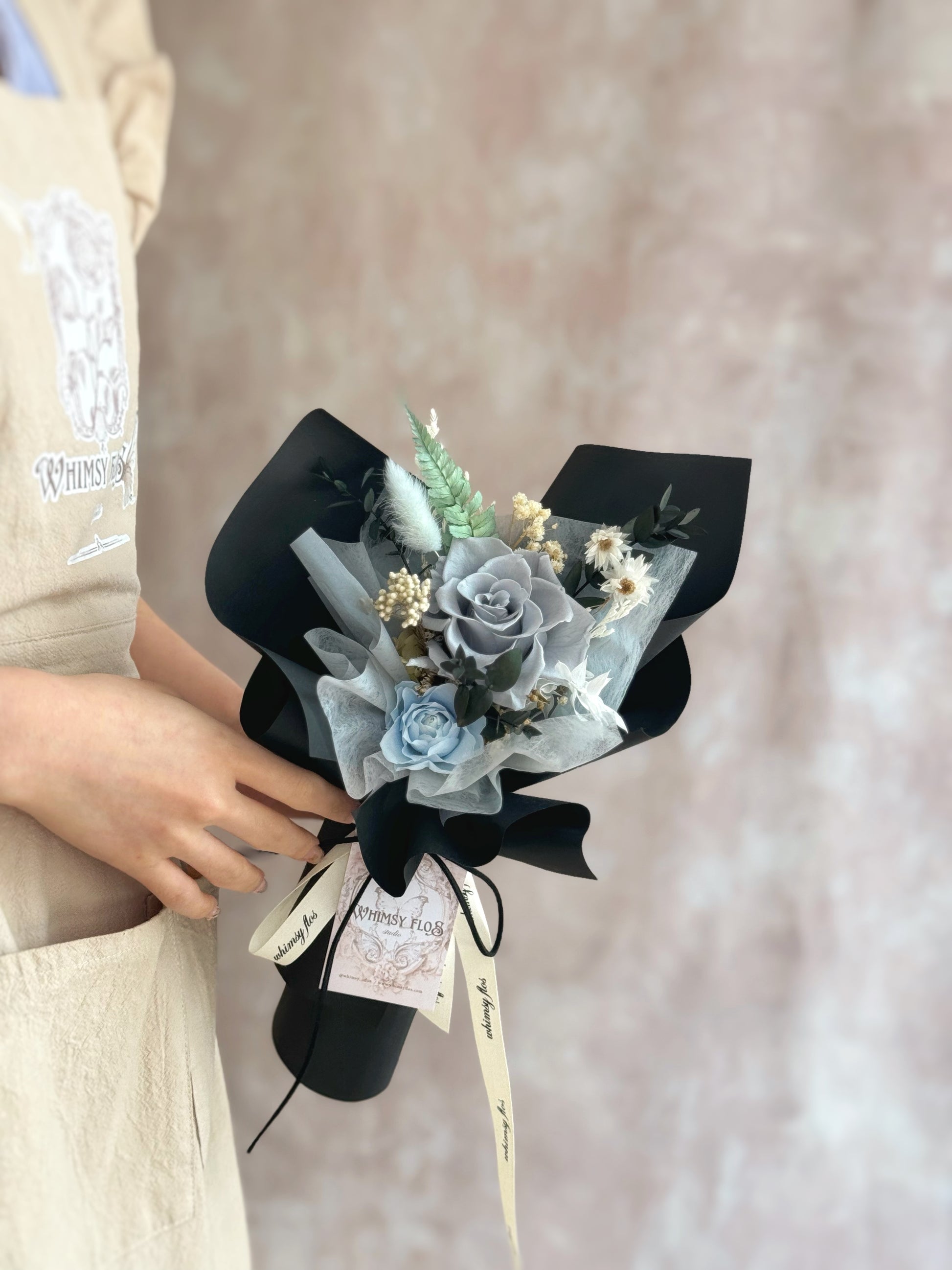 A model holding the Finn bouquet, highlighting its elegant and refined design with deep black and blue preserved roses, perfect for the modern man.
模特手持“Finn”花束，突显其优雅精致的设计，深黑与蓝色的永生玫瑰，完美契合现代男士的品味。