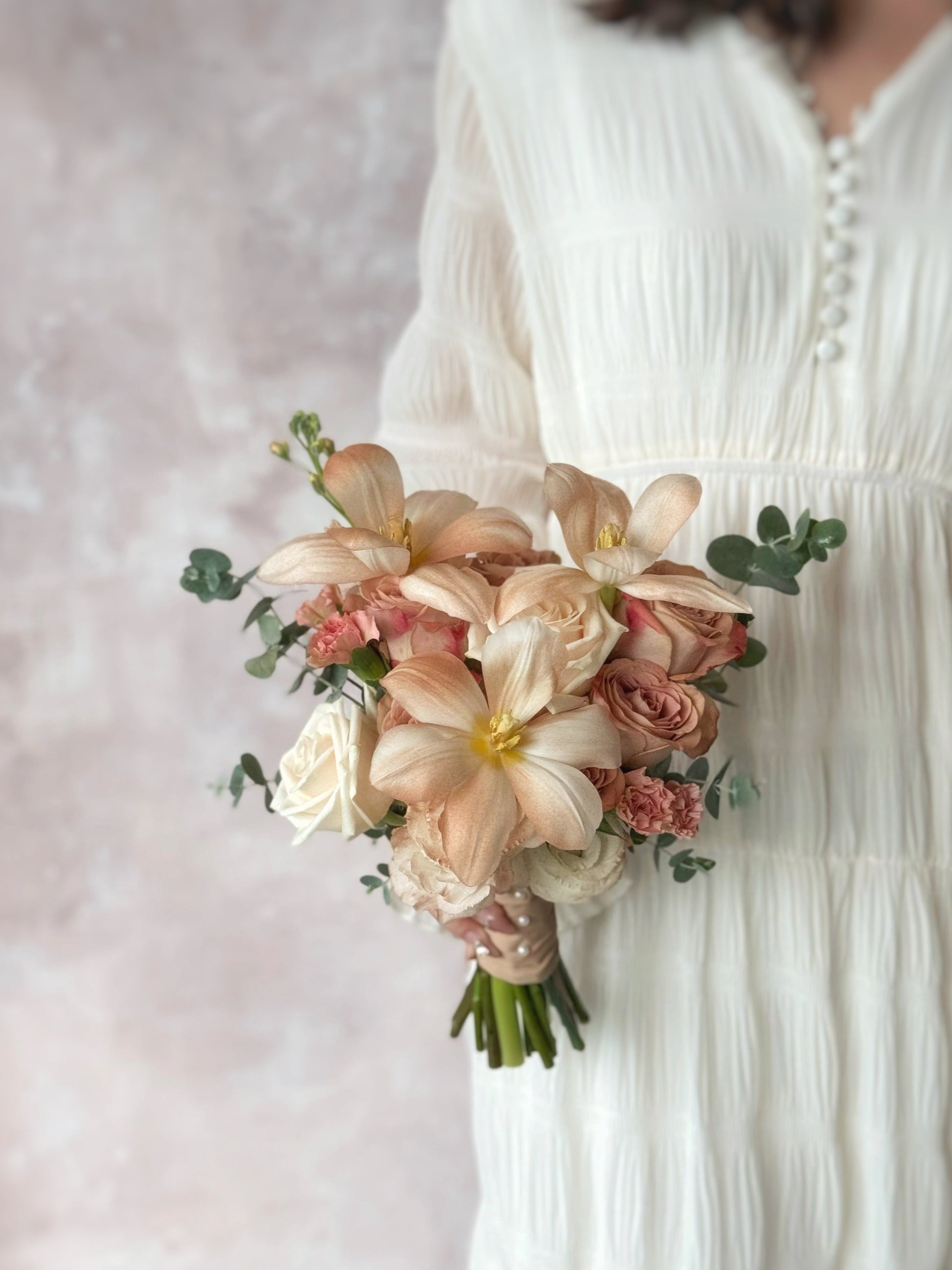 Model in white dress holding Florette best selling bridal bouquet, a muted-tone bouquet with cappuccino roses, soft pastel florals, and earthy tulips, creating a dreamy and romantic look.
新娘手持大地色调的新娘捧花，卡布奇诺玫瑰与柔和粉彩花朵相映，散发温柔浪漫气息


