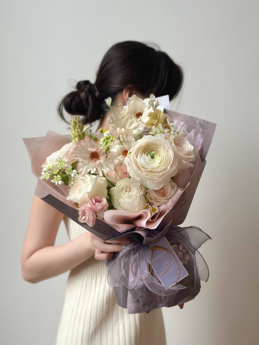 Front view of a soft cream and blush flower bouquet wrapped in dusty lavender paper, showcasing a serene and elegant look.
正面拍摄的柔和奶油色与粉色花束，搭配薰衣草灰包裹纸，展现宁静优雅的氛围。