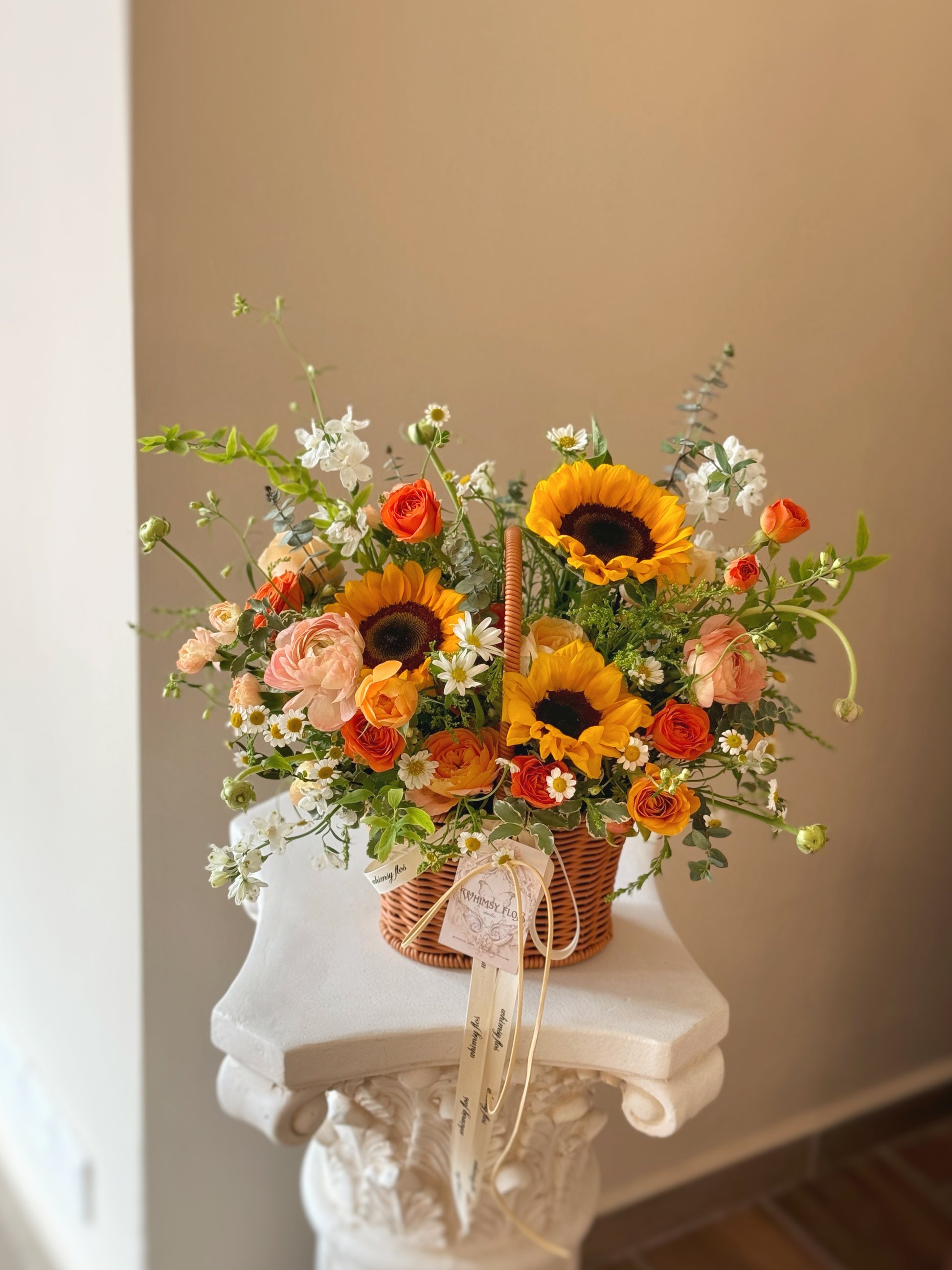 Front view of a vibrant basket arrangement with sunflowers, orange and yellow blooms, and abundant green foliage, radiating warmth and cheer.
正面展示充满阳光感的花篮，向日葵搭配橙黄花朵与繁茂绿叶，散发温暖与喜悦氛围。