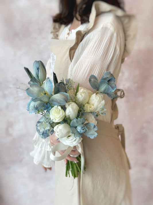 Introducing our Blue Haze fresh flowers bridal bouquet, featuring stem dyed dusty blue tulips, roses, eustoma, mini delphinium and other fillers.