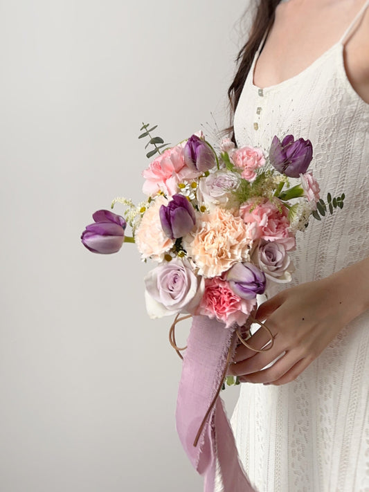  A delightful mix of purple tulips, sweet pink blooms, and soft pastel accents, creating a dreamy and romantic floral arrangement. This bouquet is perfect for brides who love a playful yet elegant touch, blending purplish hues with soft pink tones for a whimsical and charming wedding look. 
漫梦幻的新娘捧花，搭配紫色郁金香、粉色玫瑰和柔和的粉彩花卉，营造甜美温馨的婚礼氛围