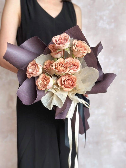 Front view of a romantic bouquet featuring cappuccino roses in dusty pink, wrapped in deep mauve sculptural layers for a soft yet dramatic look.
正面展示复古浪漫花束，焦点为尘粉色卡布奇诺玫瑰，搭配深紫包裹，整体呈现柔和却带有神秘气质的风格。