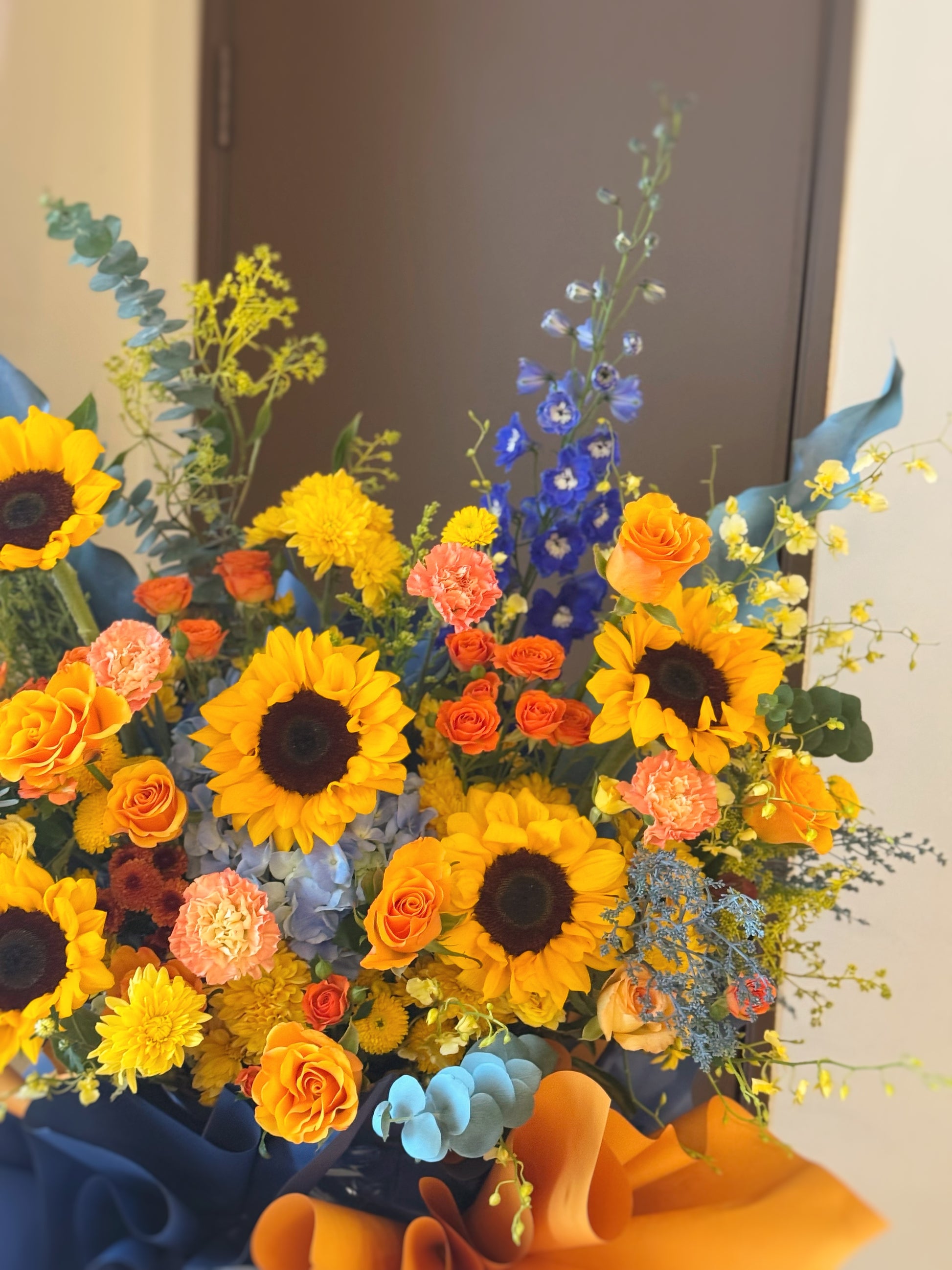 Detailed view of grand opening stand — sunflower faces, vibrant orange blooms, and blue accent flowers among greenery.
细节视角：金黄向日葵、亮橙花瓣与蓝色点缀花材在绿叶中相互映衬