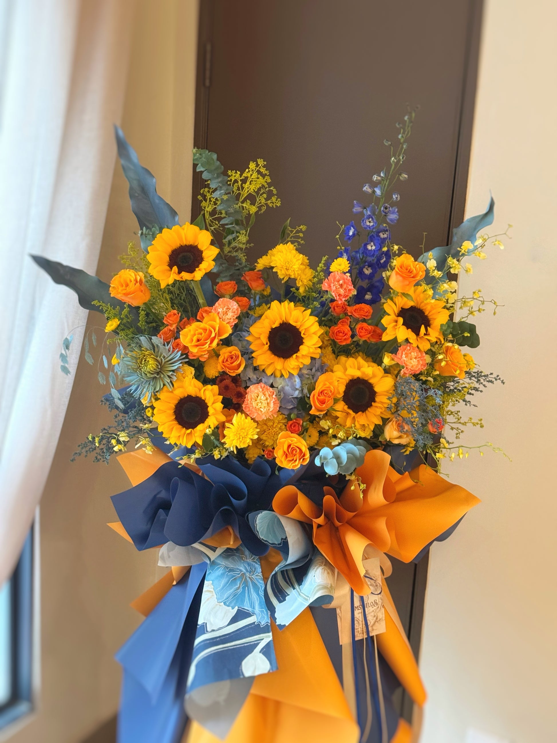 Front view of Marigold Twilight grand opening flower stand with radiant sunflowers, bright orange blooms, and blue-toned accents against lush greenery. 
明亮向日葵与鲜艳橙色花材，搭配蓝色点缀与绿叶，视觉醒目