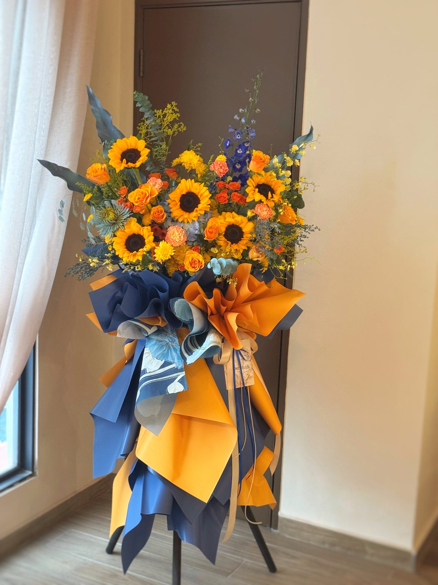 Vibrant grand opening flower stand with radiant sunflowers and orange flowers, accented by blue tones and greenery, viewed from the front.