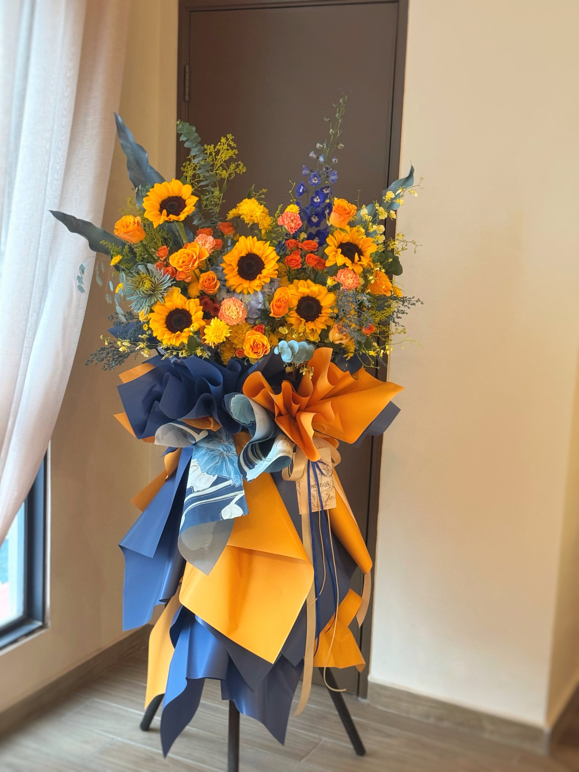 Vibrant grand opening flower stand with radiant sunflowers and orange flowers, accented by blue tones and greenery, viewed from the front.