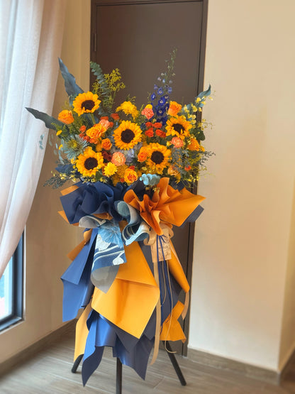 Vibrant grand opening flower stand with radiant sunflowers and orange flowers, accented by blue tones and greenery, viewed from the front.