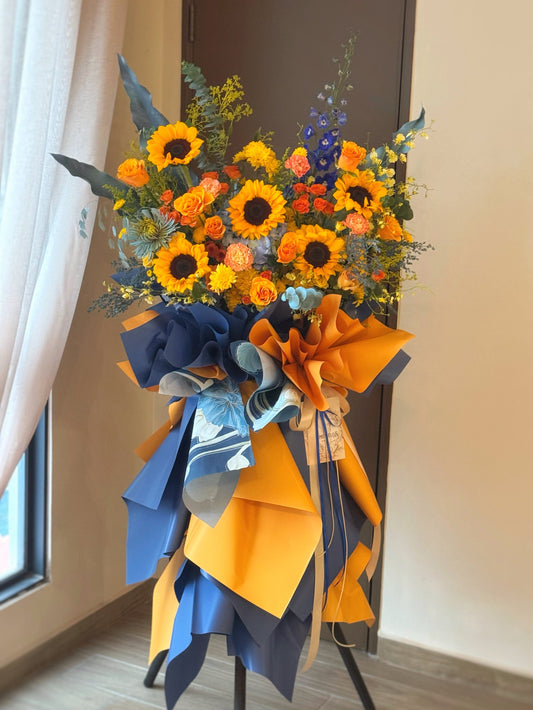 Vibrant grand opening flower stand with radiant sunflowers and orange flowers, accented by blue tones and greenery, viewed from the front.