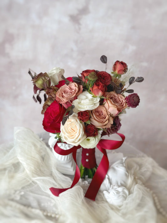 Muted-tone bridal bouquet resting on a table, showcasing a unique color palette of mauve, maroon, soft white, and dusty blush blooms
充满情绪感与戏剧性的婚礼捧花，深紫红与酒红色调与柔和的白色及浅粉花朵形成优雅对比

