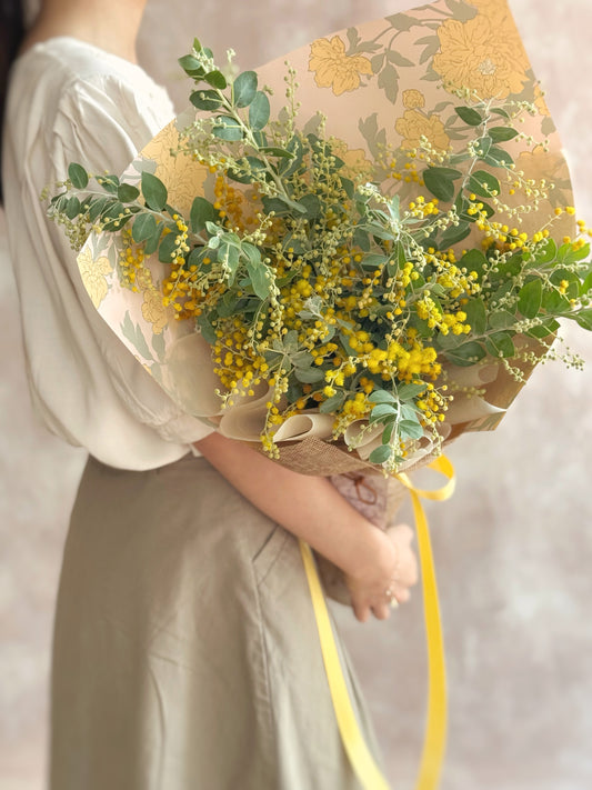 Model holding Golden Glow bouquet with sunny yellow acacia blooms, adding a cheerful touch to festive styling.
 模特手持 Golden Glow 花束，明亮黄色合金欢花，为节日布置增添阳光感。