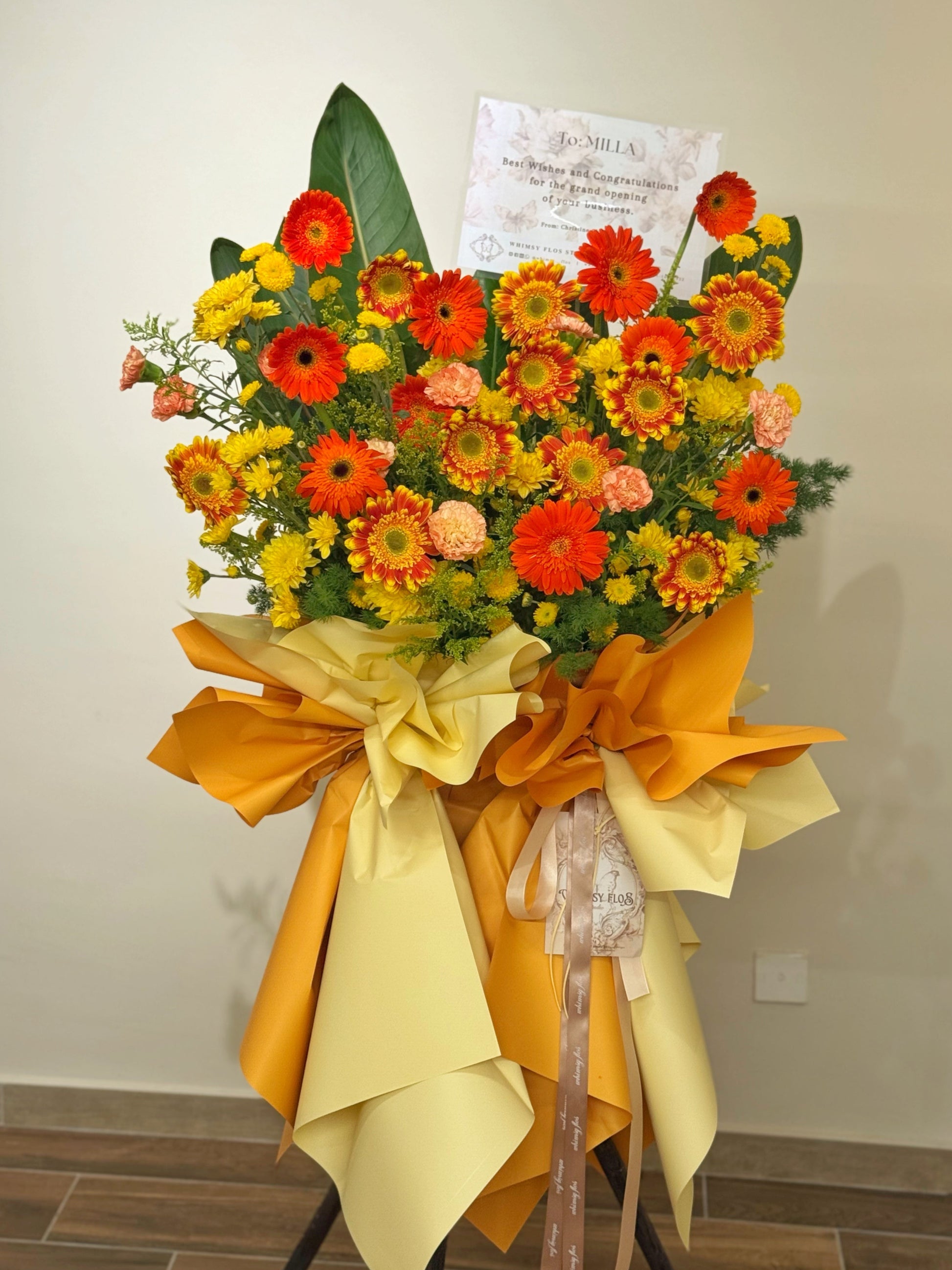 Fresh flower grand opening stand against a plain wall, featuring bright orange and sunny yellow gerberas with lush greenery, arranged to create a vibrant, energetic, and joyful display for celebrations.