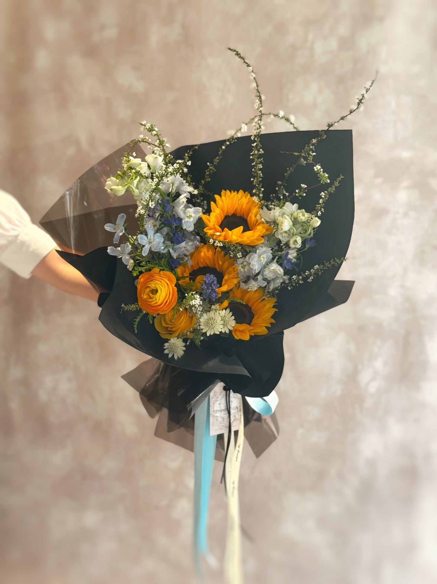Front view of a bold sunflower bouquet with bright yellow blooms paired with cool-toned blue flowers, wrapped in sleek black for striking contrast.

 正面展示个性鲜花花束，金黄向日葵搭配冷调蓝色花材，外层以黑色包装突显强烈对比感，视觉冲击力十足。
