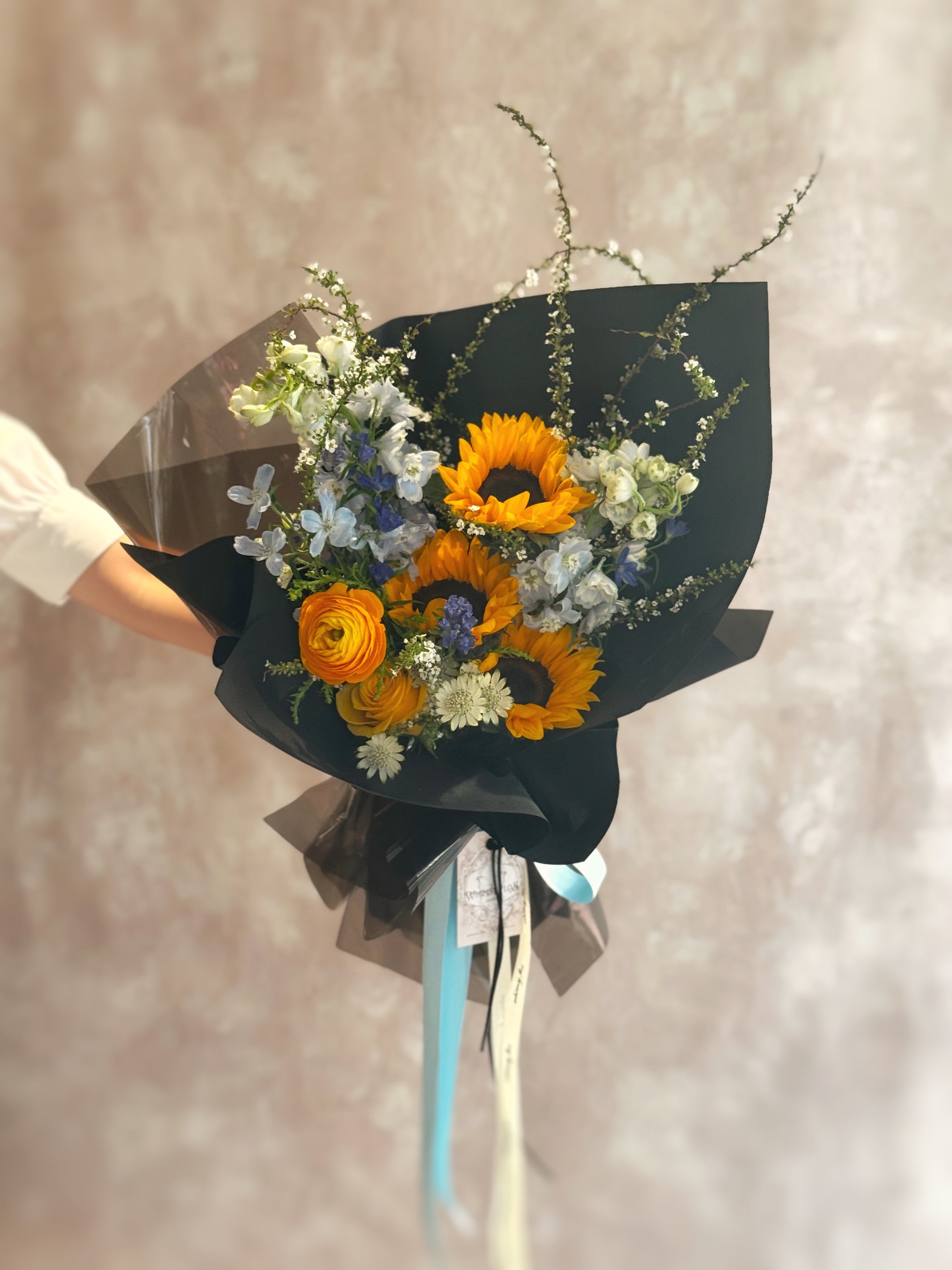 Front view of a bold sunflower bouquet with bright yellow blooms paired with cool-toned blue flowers, wrapped in sleek black for striking contrast.

 正面展示个性鲜花花束，金黄向日葵搭配冷调蓝色花材，外层以黑色包装突显强烈对比感，视觉冲击力十足。