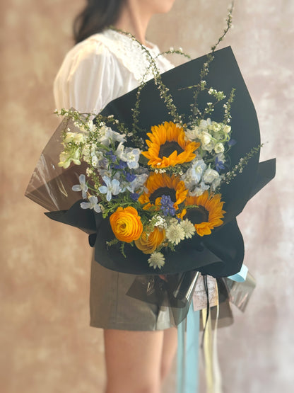 Model holding the bouquet in hand, sunflowers facing forward with the black wrapping framing the vivid yellow and blue tones — a statement piece.
模特手持花束正面展示，黑色包装衬托出向日葵与蓝色花材的亮眼组合，是引人注目的时尚花礼。