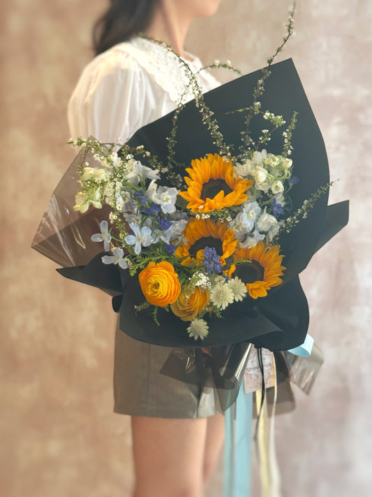 Model holding the bouquet in hand, sunflowers facing forward with the black wrapping framing the vivid yellow and blue tones — a statement piece.
模特手持花束正面展示，黑色包装衬托出向日葵与蓝色花材的亮眼组合，是引人注目的时尚花礼。