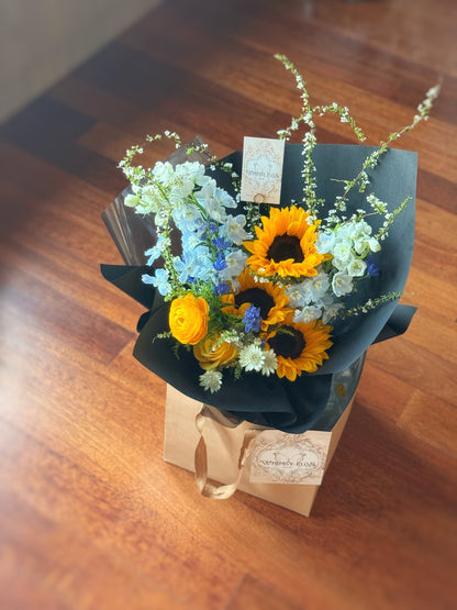 Top-down view showing bold color blocking between the golden sunflowers and the surrounding blue blooms, framed by matte black wrapping.
 俯视图呈现黄蓝花材色块拼接效果，整体包裹以哑黑色纸张收边，简约中见张力。

