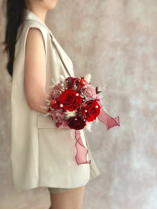 Front view of a preserved round bridal bouquet featuring nude pink, red, maroon, and cream-toned flowers with pearl accents, arranged in a classic and elegant style.
正面展示永生圆形新娘捧花，柔裸粉、浪漫红、深酒红与奶油色调交织，点缀珍珠装饰，整体风格典雅迷人。