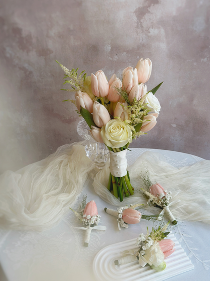Photo of Korean style peachy pink tulip bridal bouquet on a table, together with  delicate corsages featuring a single peachy tulip and white floral accents, tied with an elegant silk ribbon.
精致韩式蜜桃粉郁金香新娘捧花和与胸花，蜜桃色郁金香搭配白色花朵点缀，丝带装饰更显高贵雅致。