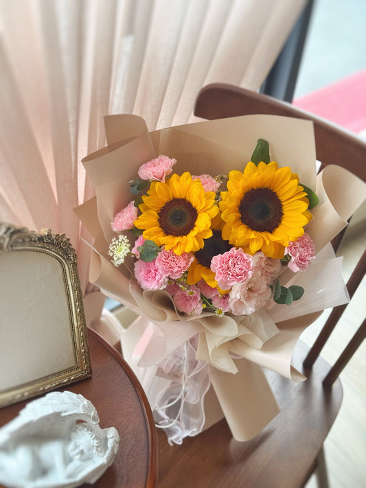 Sunflower and carnation bouquet laid on a table surface, emphasizing its fresh garden feel and rustic charm.

向日葵和粉色康乃馨花束摆放于木椅子上，散发自然花园氛围与朴实温馨的风格。