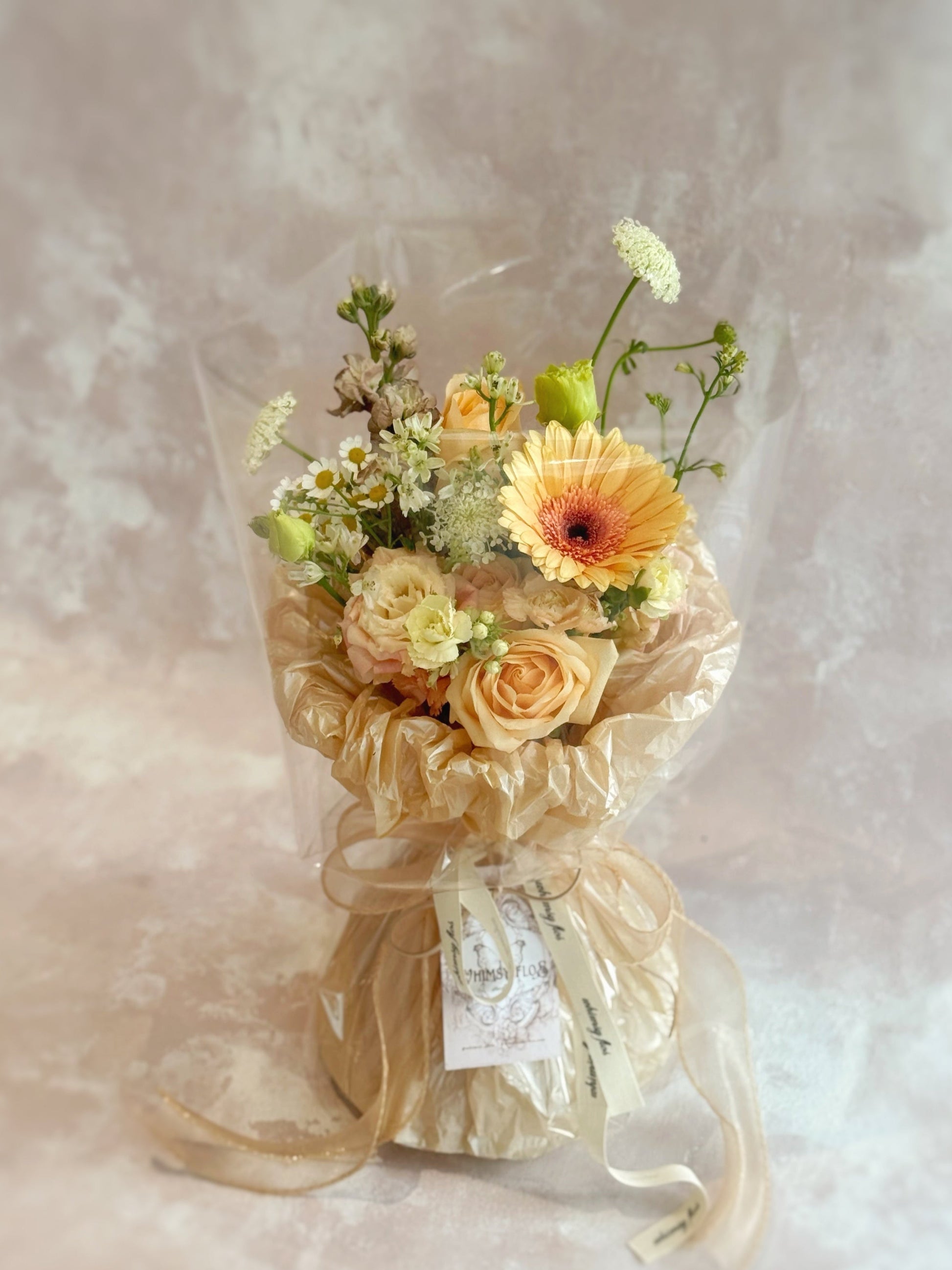 Front view of a hand-tied bouquet featuring fresh florals in peach, cream, khaki, and light brown tones, wrapped in layered textured paper and a sheer wrap.
 正面展示手绑花束，由蜜桃、奶油、卡其与浅棕色调花材组成，搭配多层纹理纸与透明轻盈外层包裹。