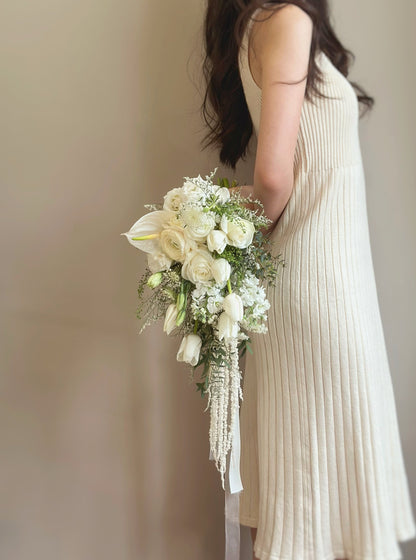 A bride elegantly holding a cascading teardrop bouquet, where white tulips and soft greenery flow gracefully to create a natural and sophisticated look. The bouquet’s elongated silhouette adds an element of movement and charm, making it a captivating choice for brides who love a fairytale aesthetic.
新娘优雅地手持一款垂坠式水滴形捧花，白色郁金香与柔和绿植轻盈流动，营造出自然且高贵的氛围。修长的花束轮廓增添了动感和迷人魅力，非常适合喜爱仙境风格的新娘。